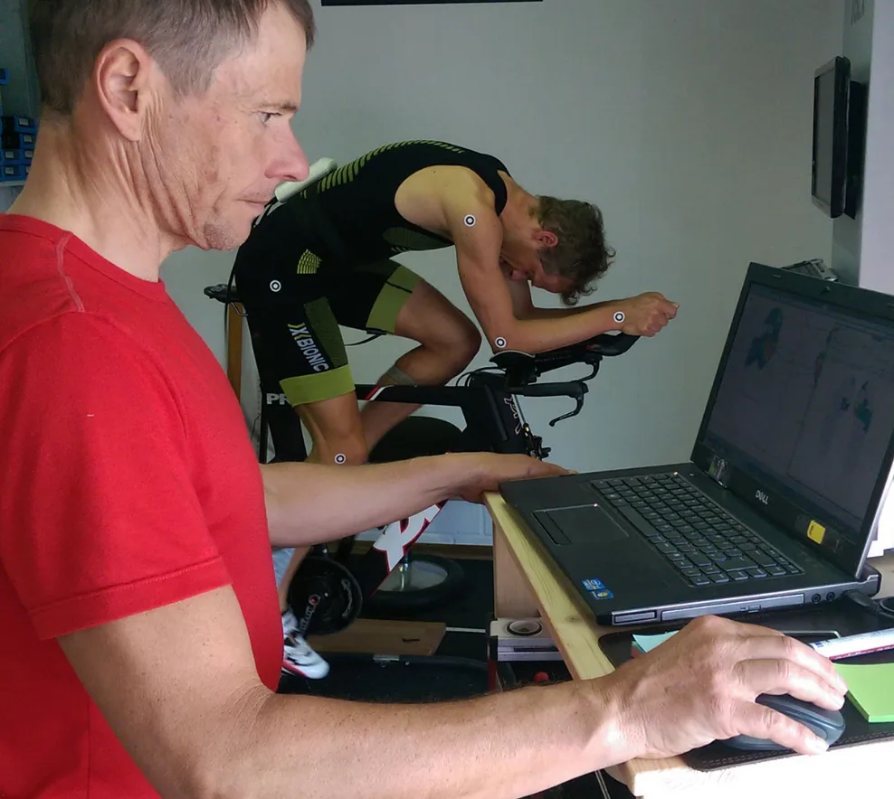 Trainer überprüft Daten am Laptop während Radprofi in Aeroposition auf Rennrad-Trainer fährt (Bikefitting, Leistungsdiagnostik)